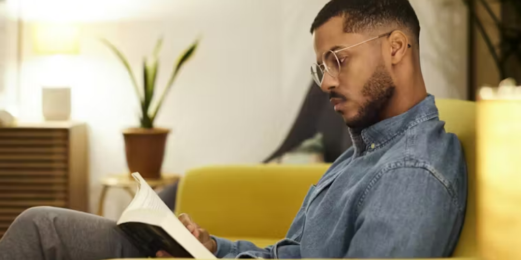 A brown-skinned man wearing glasses sits in a comfy yellow chair reading a book. (Credit: Morsa Images/DigitalVision via Getty Images)